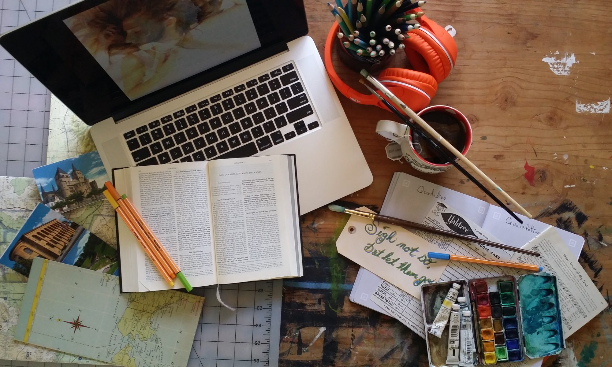 A table scape showing headphones, a journal, bible, art supplies, maps, a laptop, tea, and game cards.